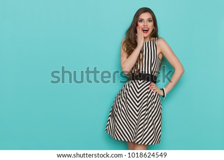 Surprised beautiful young woman in elegant black and white stroped cocktail dress is holding hand on chin, shouting and looking at camera. Three quarter length studio shot on turquoise background.