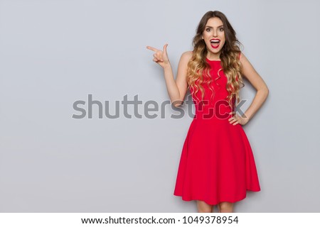 Surprised beautiful young woman in red dress is pointing and shouting. Three quarter length studio shot on gray background.