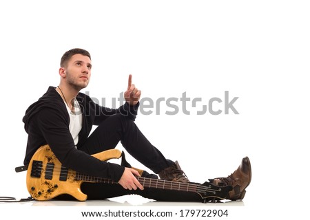 Male guitarist with bass guitar sitting on the floor and pointing up at copy space. Full length studio shot isolated on white.