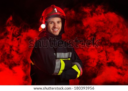 Fireman in red helmet posing with arms crossed. Three quarter length studio shot on black background with red smoke.