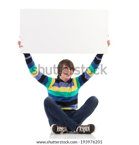 Smiling young man sitting with legs crossed and holding white banner over his head. Full length studio shot isolated on white.