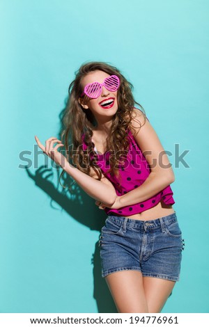 Smiling young woman posing in heart shaped glasses. Three quarter length studio shot on teal background.