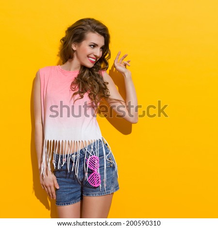 Flirting girl. Smiling beautiful young woman is waving hand and looking away. Three quarter length studio shot on yellow background.