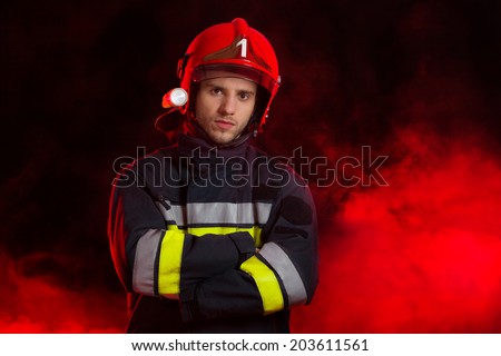 Portrait of the fireman. Serious fireman in red helmet standing with arms crossed. Waist up studio shot on black background and red smoke.