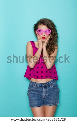 Beautiful young woman in sunglasses looking surprised. Three quarter length studio shot on teal background.