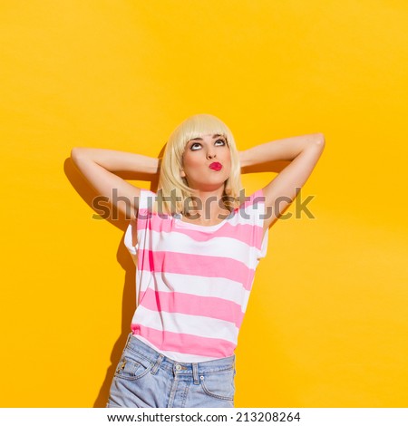 What to do? Thinking blond young woman leaning on the wall with hands behind head and looking up. Waist up studio shot on yellow background.