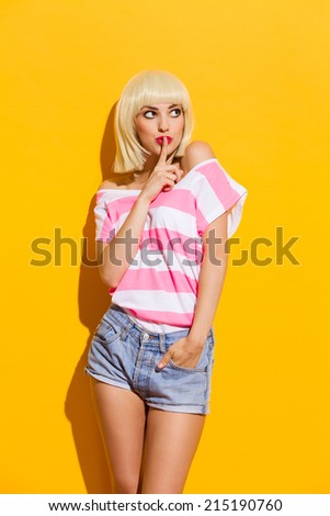 Keep it secret. Beautiful blonde young woman holding finger on her lips and looking away. Three quarter length studio shot on yellow background.