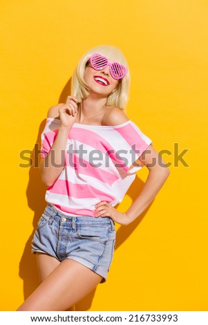 Smiling summer girl in pink glasses. Beautiful blond girl in pink sunglasses smiling. Three quarter length studio shot on yellow background.