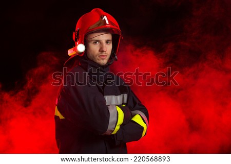 Portrait of the fireman. Serious fireman in red helmet standing with arms crossed. Waist up studio shot on black background and red smoke.