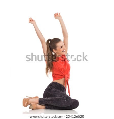 Finally weekend. Beautiful woman in red top, black jeans and high heels sitting on the floor with arms raised and shouting. Full length studio shot isolated on white.