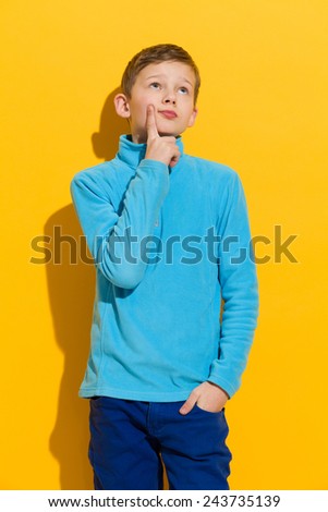 Thinking young boy standing and holding index finger on a chin. Three quarter length length studio shot on yellow background.