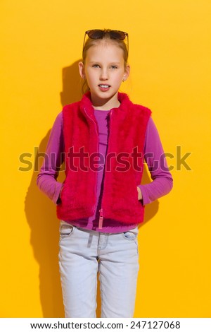 Cheerful blond girl in pink fur vest posing with hands in pockets. Three quarter length studio shot on yellow background.