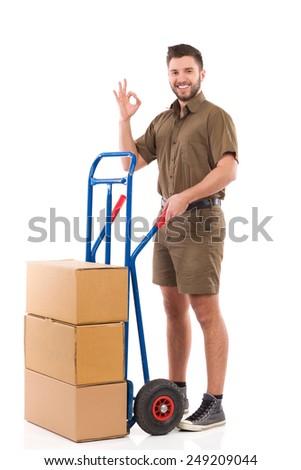 Cheerful delivery man standing with a push cart and showing ok sign. Full length studio shot isolated on white.