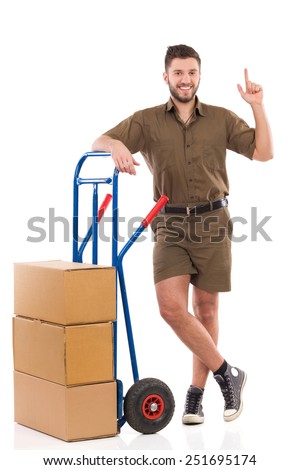 Smiling delivery man standing relaxed with a push cart and pointing up. Full length studio shot isolated on white.