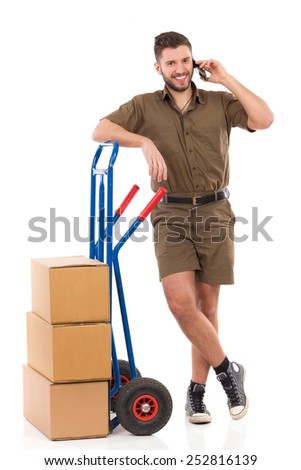 Ready for delivery. Smiling messenger standing with a push cart and using a cell phone. Full length studio shot isolated on white.