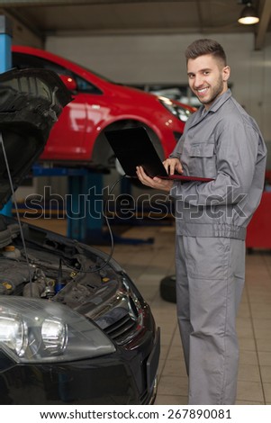 Car engine inspection. Smiling auto mechanic holding a computer connected to a car engine