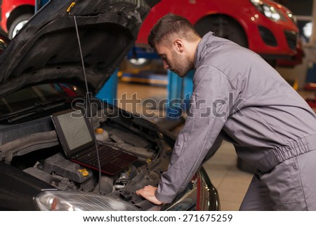 Monitor the Operation of the Engine. Focused auto mechanic looking on a computer connected to a car engine. Waist up shot in auto repair shop.