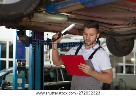 Auto Mechanic Check List. Focused mechanic standing under the car with a lamp, holding clipboard and reading.