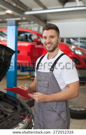 Car Warranty Review. Smiling mechanic in auto repair shop posing with a clipboard. Waist up shot against red car on a lift.