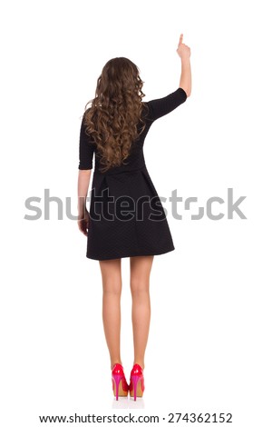 This is your copy space. Rear view of pointing girl in black short dress red high heels. Full length studio shot isolated on white.