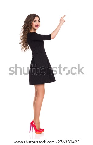 Side view of pointing beautiful elegance young woman in black mini dress and red high heels. Full length studio shot isolated on white.