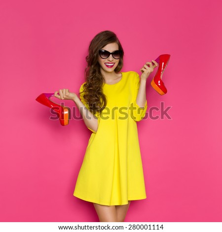 Woman And High Heels. Smiling young woman in yellow mini dress and sunglasses holding two red high heels. Three quarter length studio on pink background.