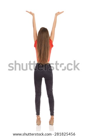 Rear view of slim young girl standing in red top, black jeans and high heels with arms raised. Full length studio shot isolated on white.