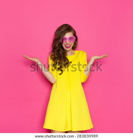 Smiling beautiful girl in yellow mini dress posing with arms outstretched and presenting something. Three quarter length studio shot on pink background.