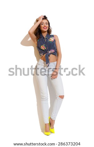 Smiling young woman in torn jeans leaning on white wall in the sunlight. Full length studio shot on white background.