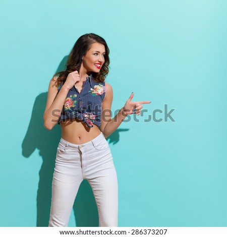 Pointing Woman On Turquoise Background. Smiling young woman pointing and looking away. Three quarter length studio shot.