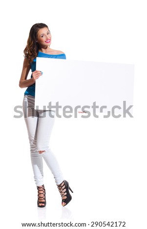 Smiling young woman in torn jeans and pink shirt standing and holding white placard. Full length studio shot isolated on white