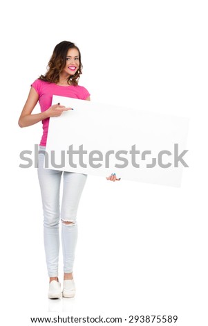 Smiling young woman in torn jeans and pink shirt standing, holding white placard and pointing. Full length studio shot isolated on white
