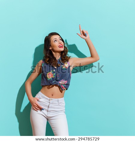 Smiling young woman pointing up and looking away. Three quarter length studio shot on teal background.