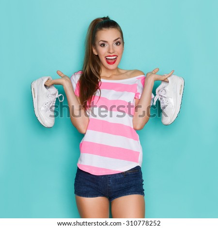 I Like Comfortable Sneakers. Shouting young woman in pink striped shirt and jeans shorts standing with arms raised and holding white sports shoes. Three quarter length studio shot on teal background.