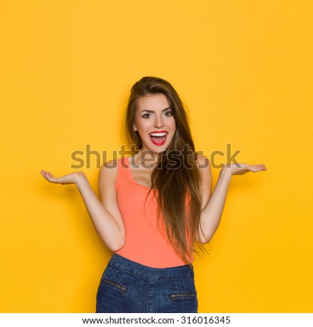 Excited Young Woman. Shouting young woman in orange shirt and jeans shorts posing with arms outstretched. Three quarter length studio shot on yellow background.