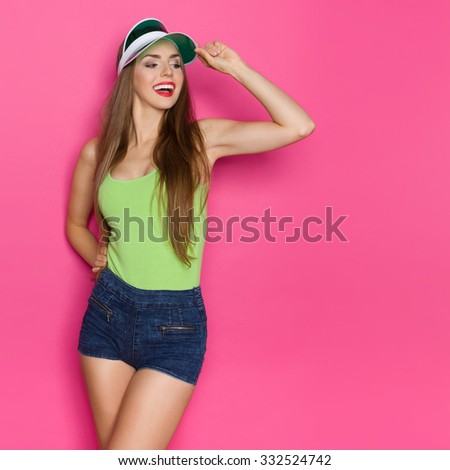 Curious Girl Looking Away. Beautiful young woman in lime green shirt, sun visor and jeans shorts looking away. Three quarter length studio shot on pink background.