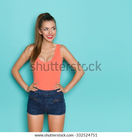 Sexy young woman in orange shirt and jeans shorts standing with hands on hip, smiling and looking away. Three quarter length studio shot on teal background.