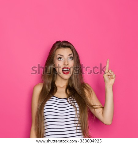 Surprised beautiful young woman in striped shirt looking up and pointing. Waist up studio shot on pink background.