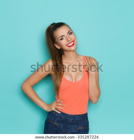 Sexy young woman in orange shirt with cleavage posing with hand on shoulder. Three quarter length studio shot on teal background.