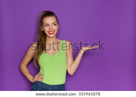 Sexy Girl Showing Product. Smiling beautiful young woman in lime green shirt and jeans shorts holding open hand and looking at copy space. Waist up studio shot on violet background.