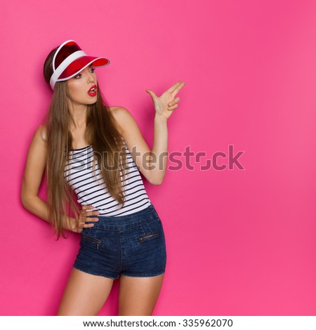 Shooting Sexy Girl. Young woman in striped shirt and red sun visor showing finger gun. Three quarter length studio shot on pink background.