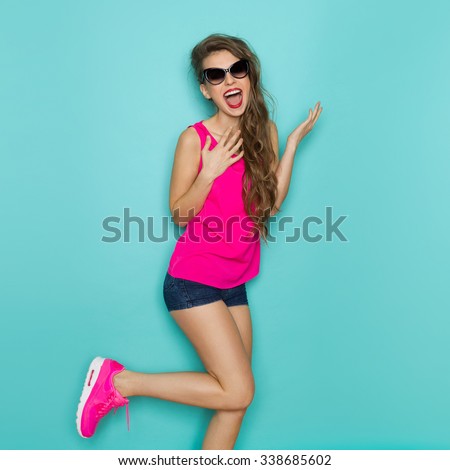 Shouting young woman in sunglasses, pink shirt, jeans shorts, and pink sneakers posing on one leg. Three quarter length studio shot on teal background.