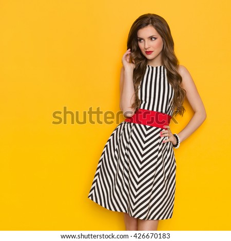 Beautiful young woman in black and white striped dress posing with hand on hip and looking away. Three quarter length studio shot on yellow background.