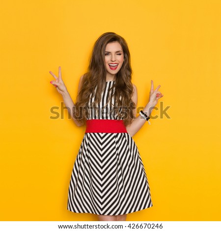 Shouting young woman in elegant striped dress winking and showing two finger or peace sign. Three quarter length studio shot on yellow background.