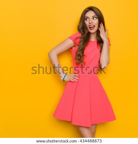 Surprised beautiful young woman with curly long brown hair standing with hand on hip and shouting. Three quarter length studio shot on yellow background.