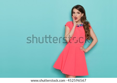 Surprised beautiful young woman in pink mini dress posing with hand on chin and looking at camera. Three quarter length studio shot on turquoise background.