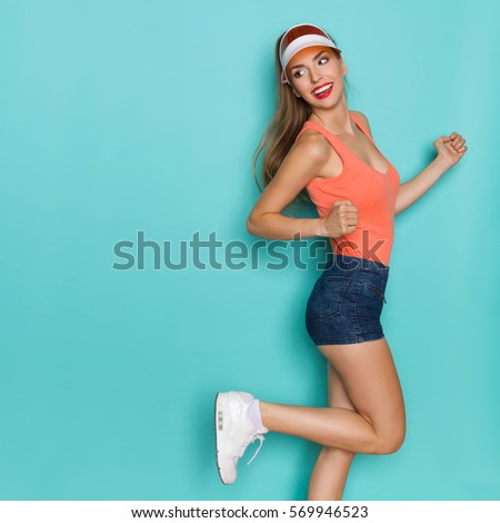 Beautiful woman in orange shirt, jeans shorts, white sneakers and sun visor standing on one leg, looking over shoulder and smiling. Three quarter length studio shot on teal background.