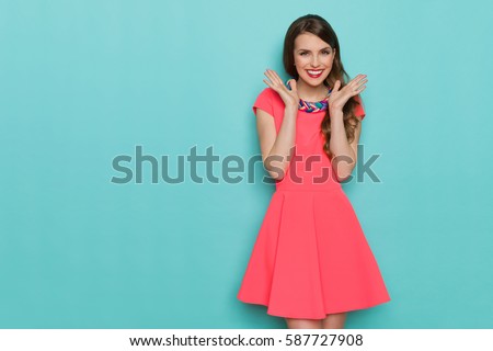 Smiling beautiful young woman in pink mini dress posing with hands on chin. Three quarter length studio shot on turquoise background.
