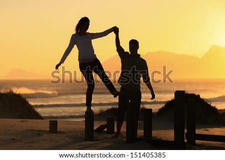 Young man helping his lady while she balances on a pillar at sunset on the beach.