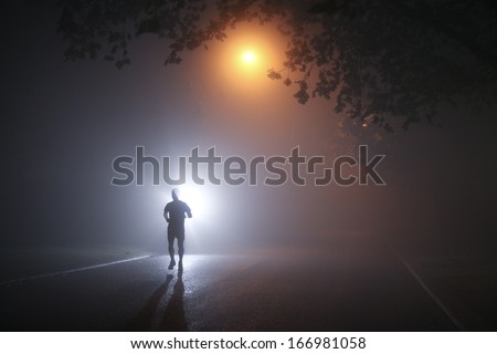 Runner Running On A Dark Misty Night Through Tree Lined Streets, Silhouetted By Street Lights.
